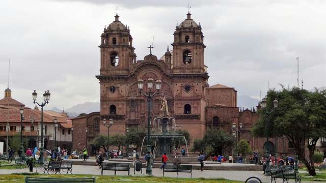 Foto de cusco com uma praça e um edifício de concreto marrom ao fundo.