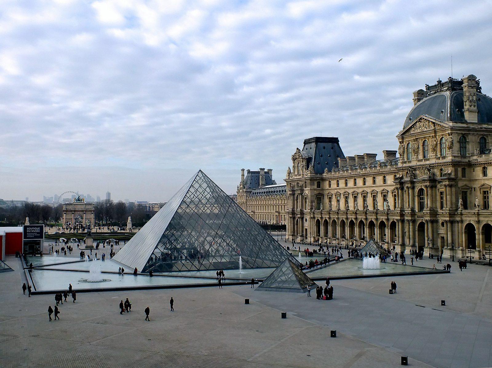 Foto do Louvre. Estrutura triangular de vidro e edificio marrom ao lado