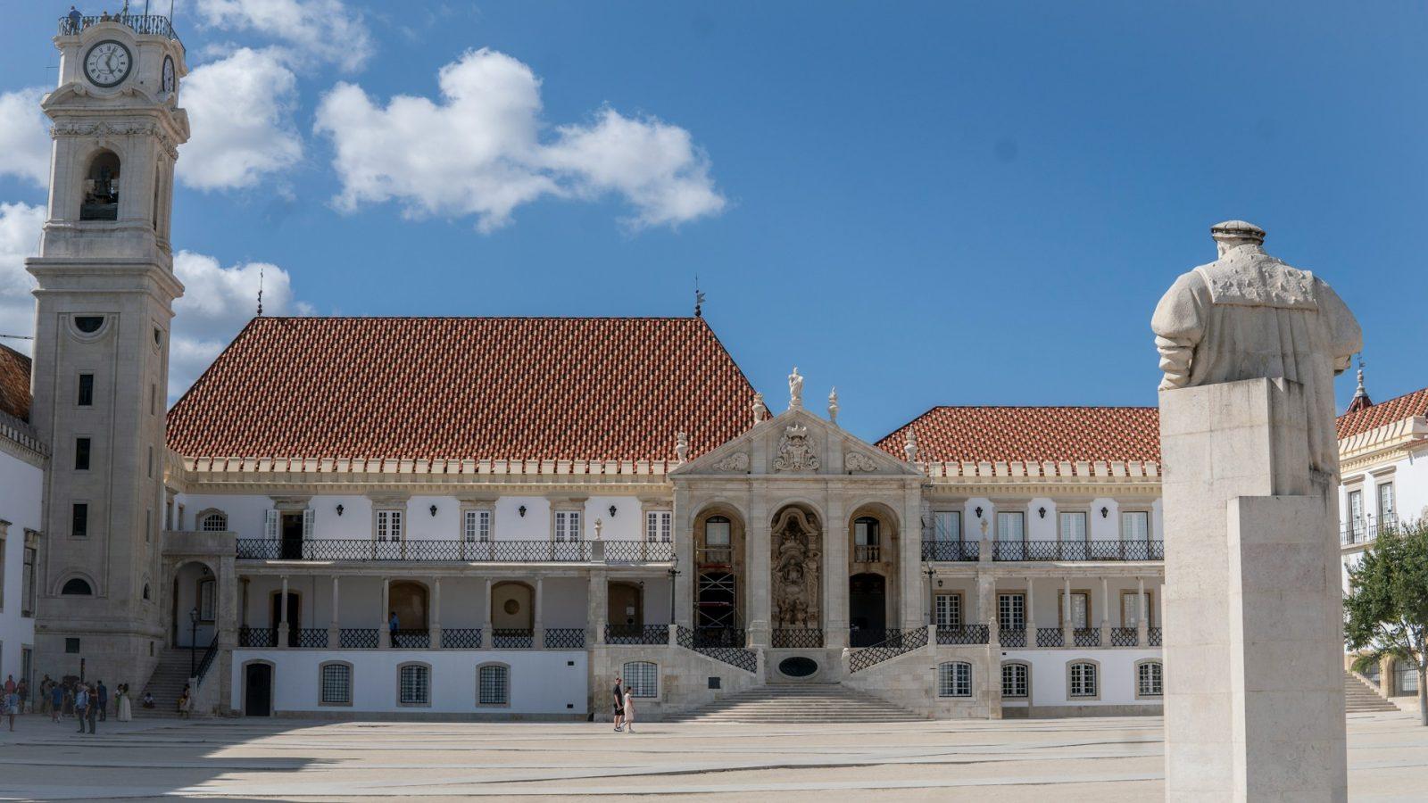 Foto da universidade de Coimbra. Escultura de concreto com grande pátio e edifício branco ao fundo.