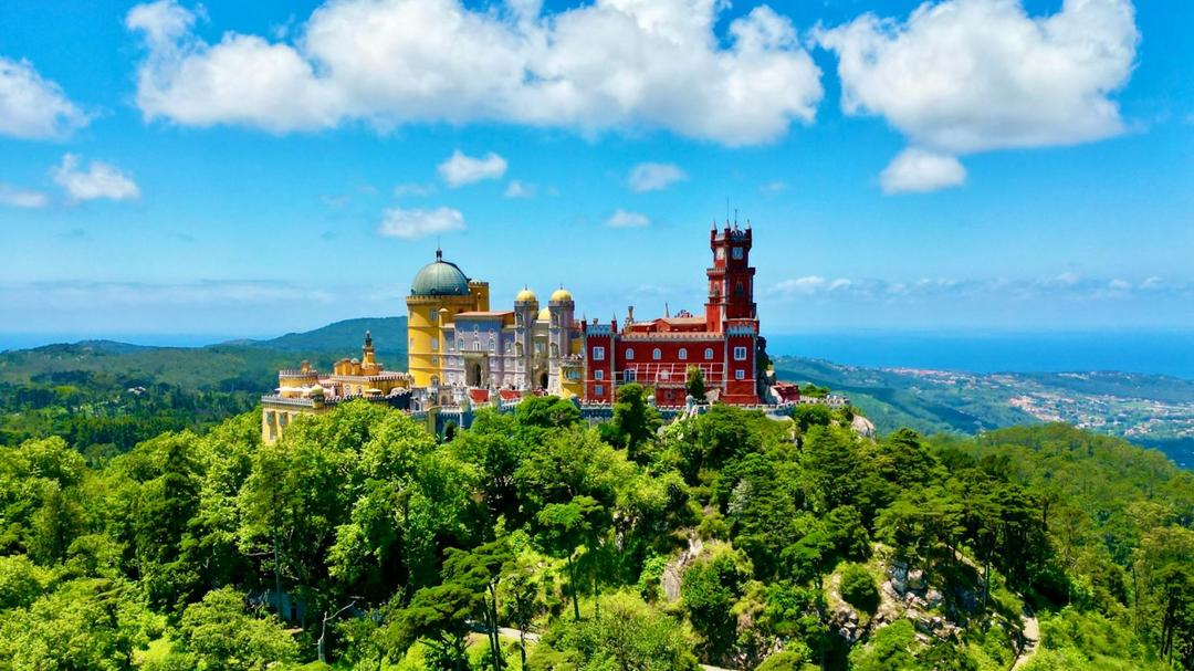 Foto de castelo em Sintra, Portugal. Castelo amarelo e vermelho rodeado por arvores verdes.